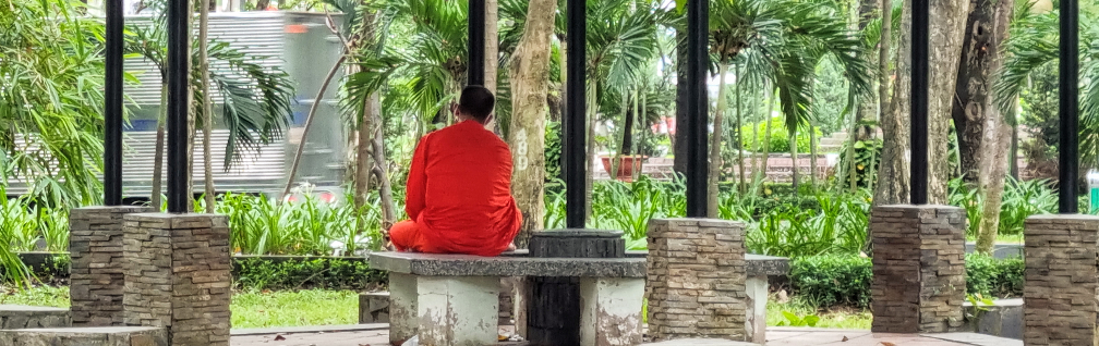Monk in orange robe sitting on park bench seemingly meditating but actually checking mobile phone